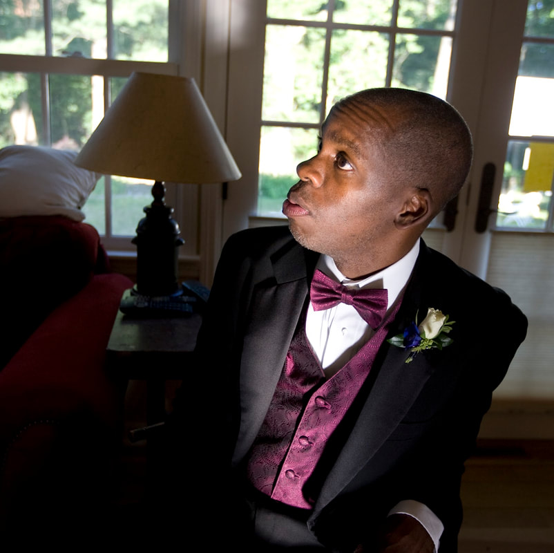 Leroy, a dark skinned Black man is seated while looking off to the side while wearing a black tux with a velvet maroon vest and matching bowtie. 