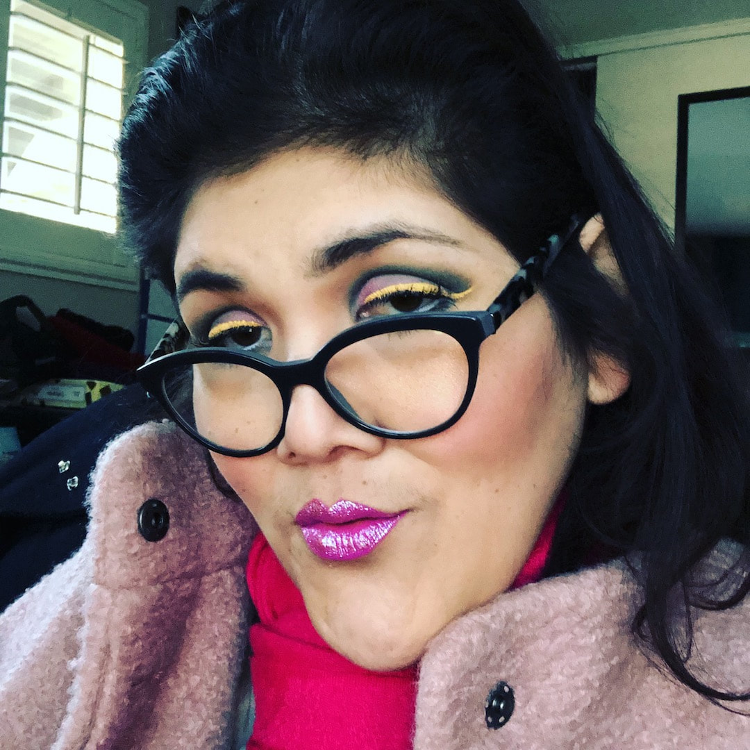 A Latinx, trans-femme looks at the camera seductively. She is wearing a pink scarf underneath a furry pink jacket. She is wearing black cat eye glasses and colorful makeup. 