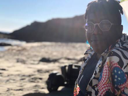 Bri smiles at the camera while wearing a multicolored shirt, hexagonal shaped purple glasses and blue glass earrings. In the background is sand, ocean and jagged cliffs. 