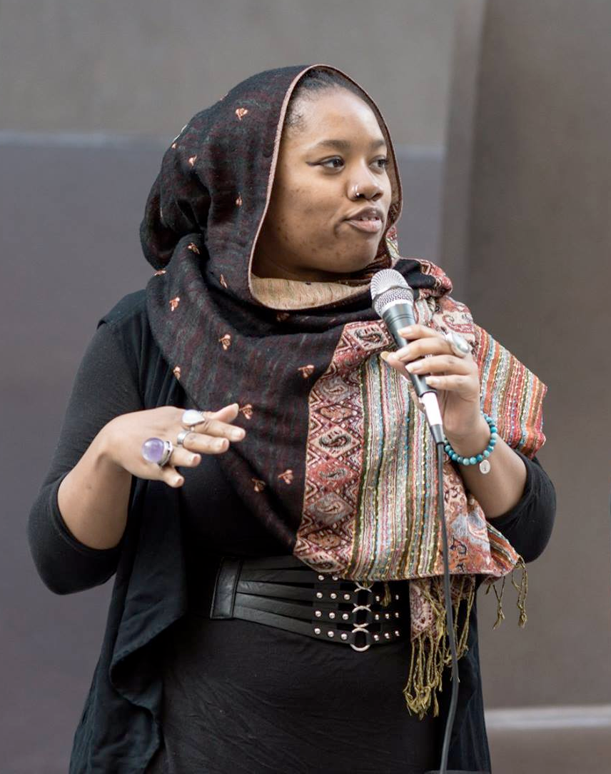 Zaynab, a Black fat woman holds a microphone to her mouth. She is wearing a black headscarf with multicolored spots and stripes.